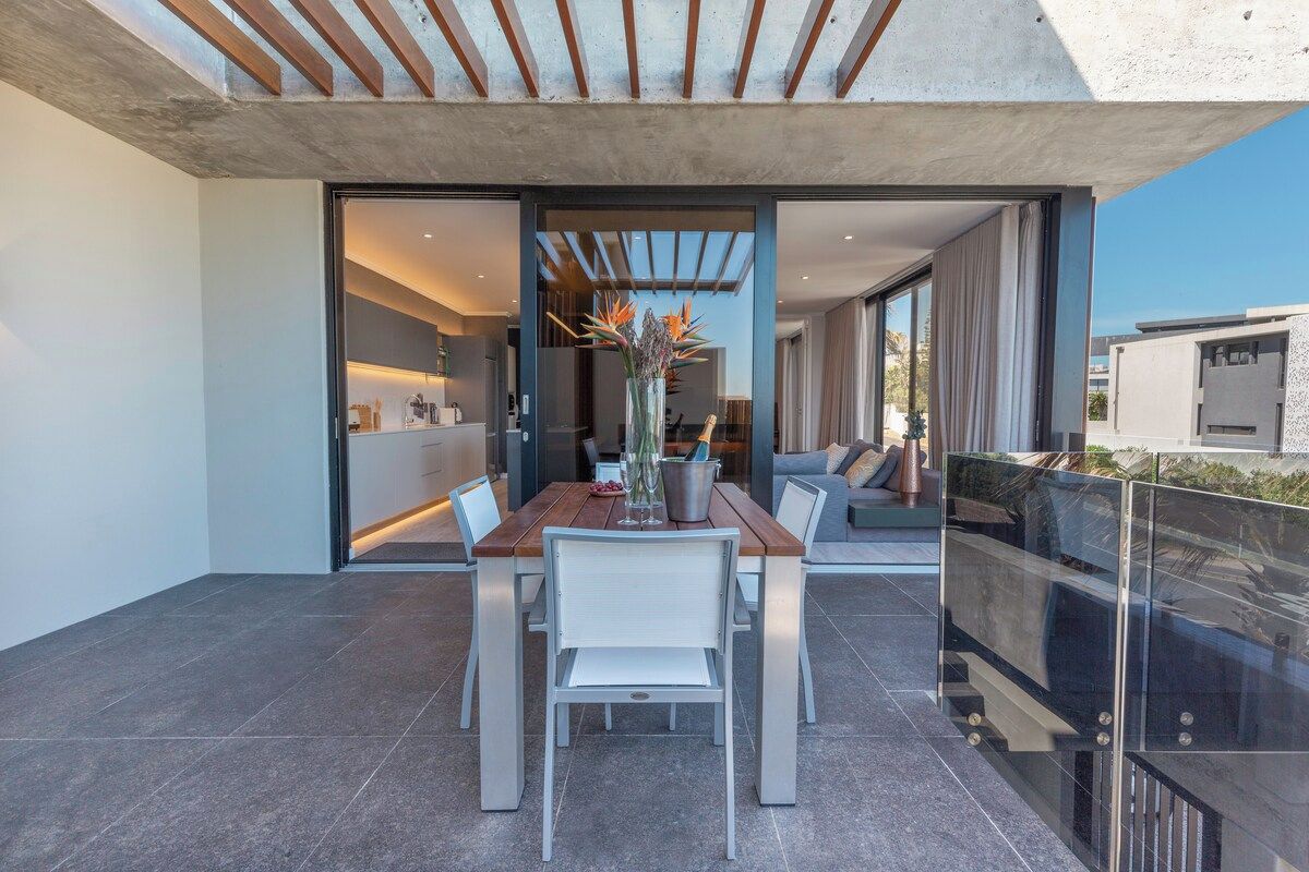 An open-plan living area with a dining table and chairs, a sofa, and a glass railing overlooking a balcony.