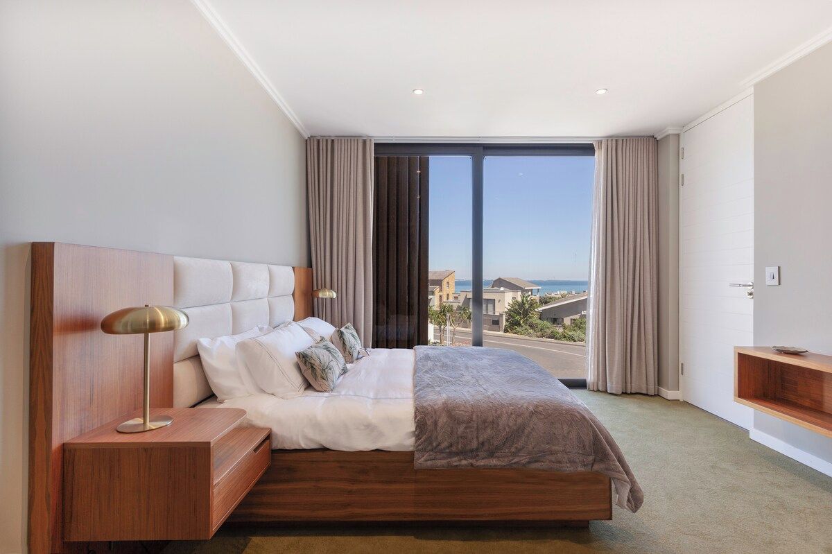 A modern hotel room with a large bed, a nightstand, a bedside table, and a view of the cityscape.