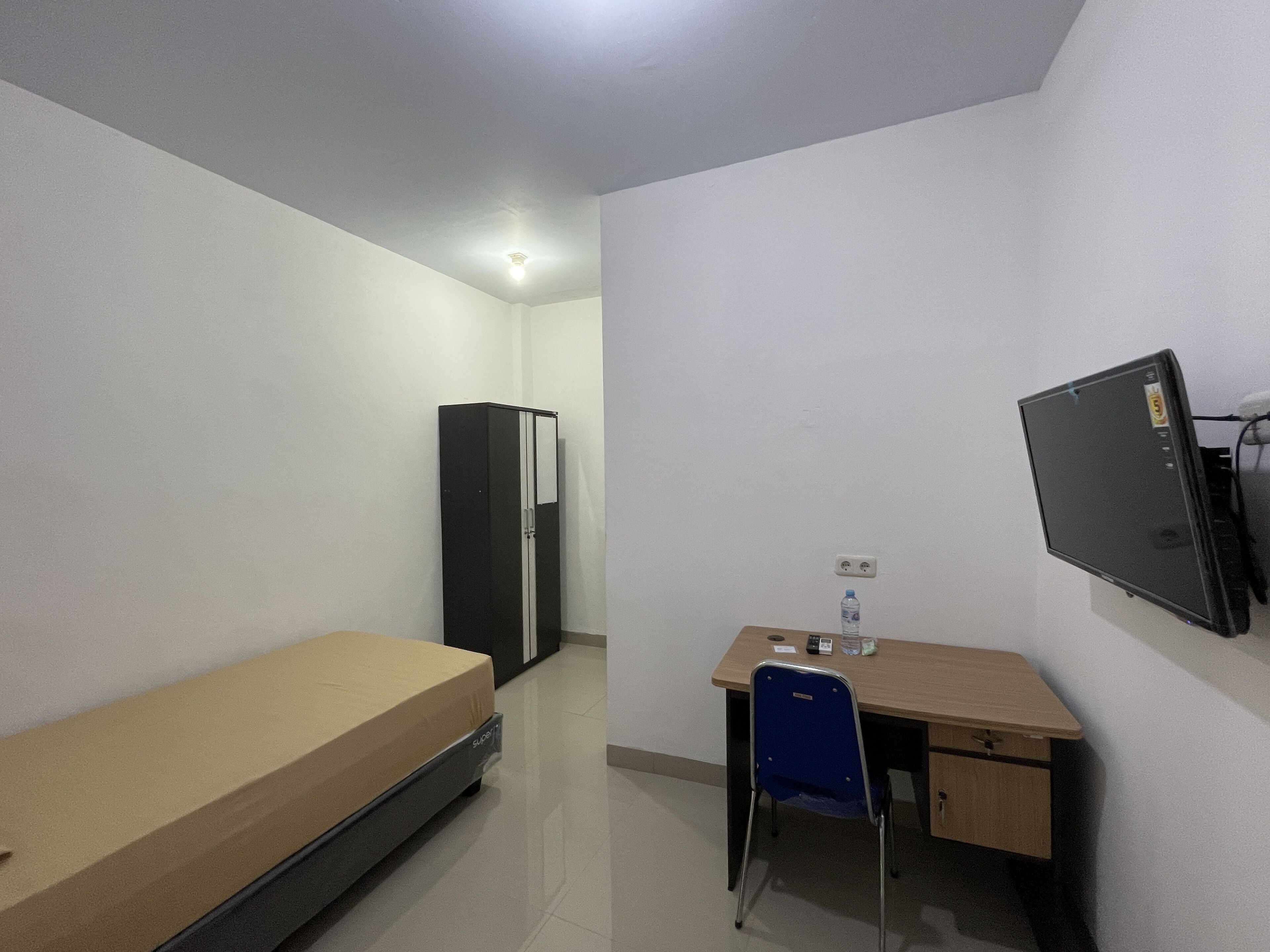A small, sparsely furnished room with a single bed, a desk with a chair, a black wardrobe, and a mounted television.