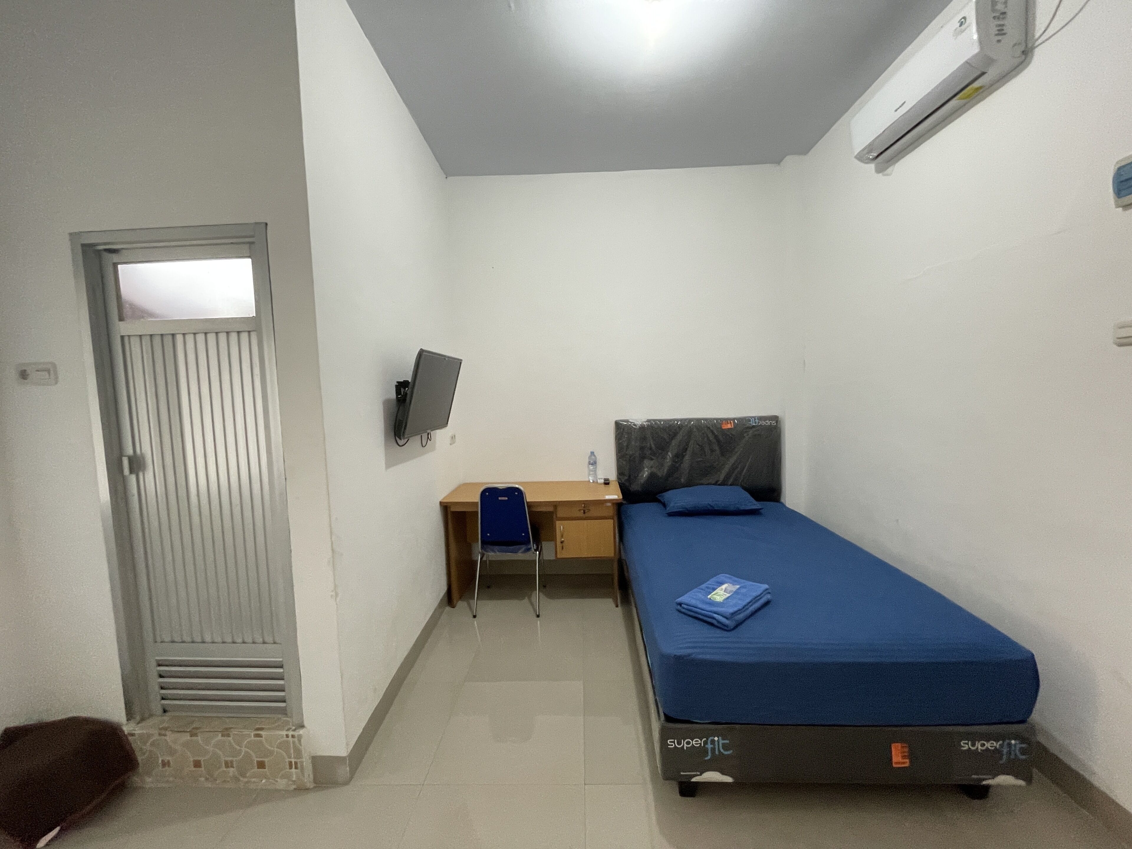 A small, well-lit room with a bed, a desk, and a chair. There is a door with a window, an air conditioner, and a wall-mounted TV.