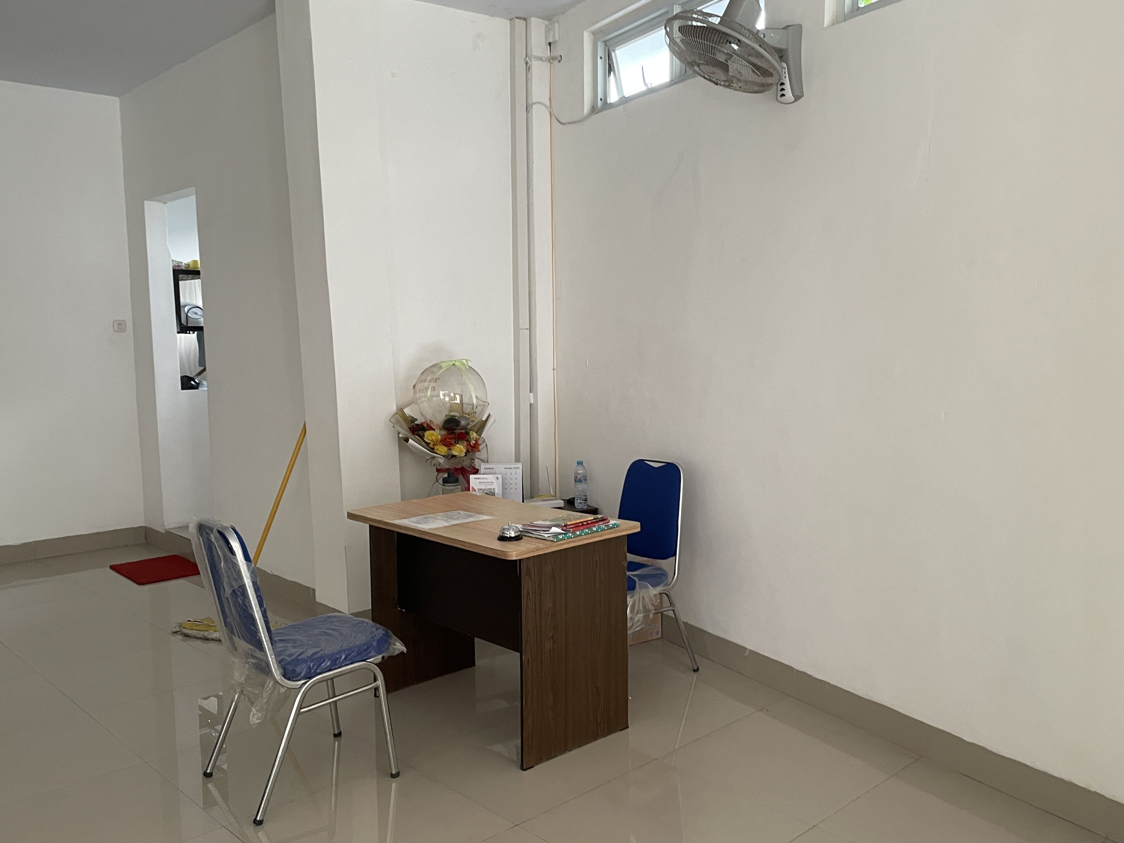 A room with a wooden desk, a chair, a standing fan, a water bottle, and a bouquet of flowers.