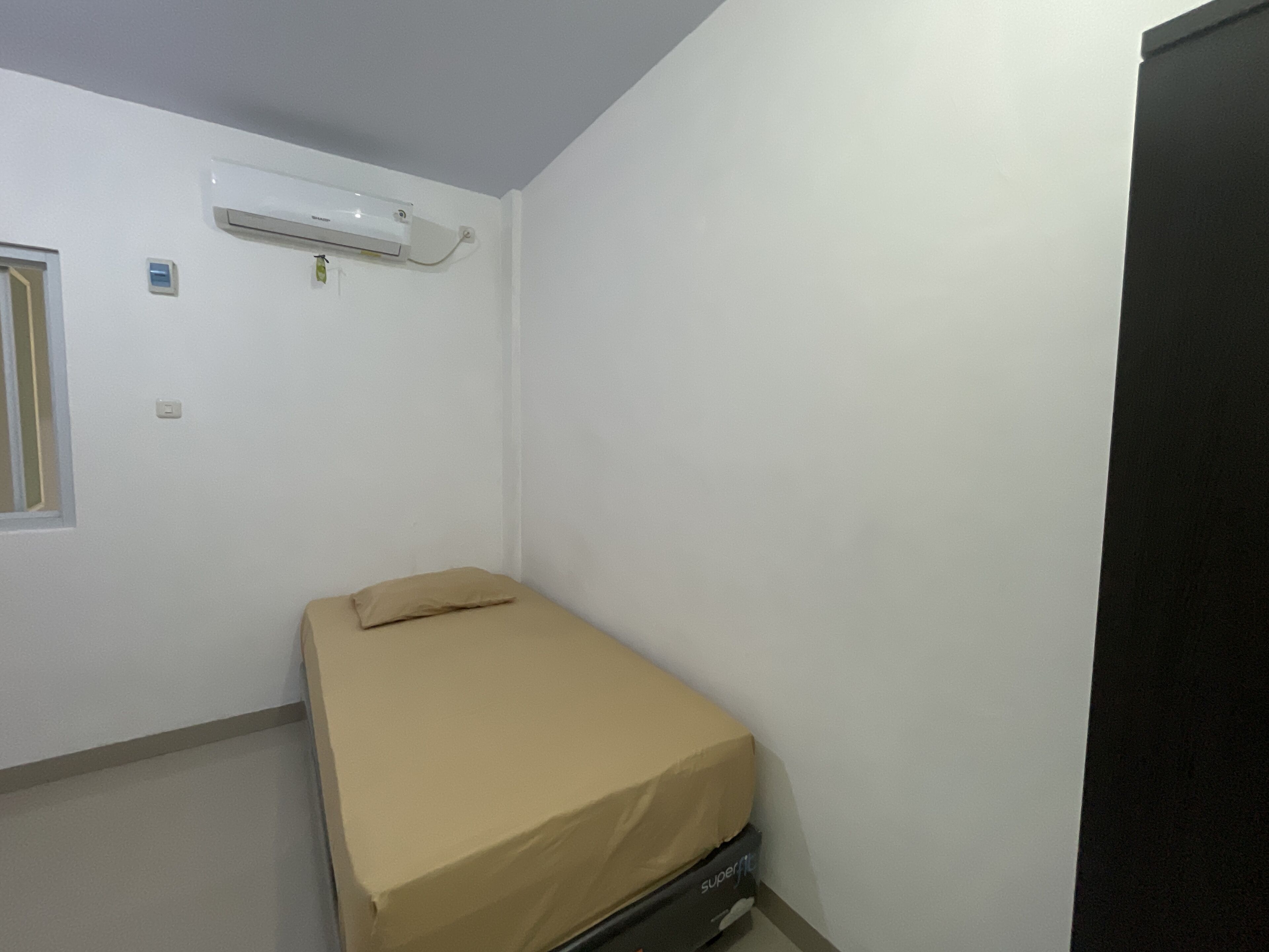 A small, single-bed room with a beige mattress, a black wardrobe, and a white air conditioning unit on the wall.