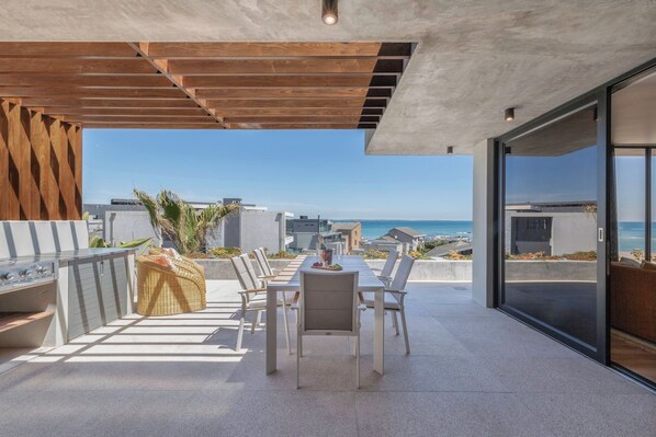 An outdoor patio with a dining table and chairs, a grill, and a view of the ocean. - State of the Art Bloubergstrand Breakaway! (Cape Town)