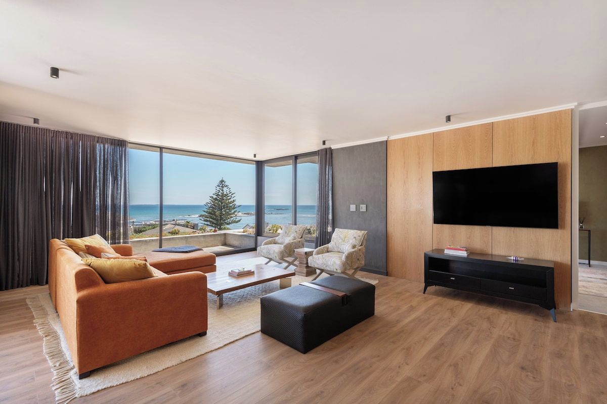 A modern living room with a large TV, comfortable seating, and a view of the ocean.