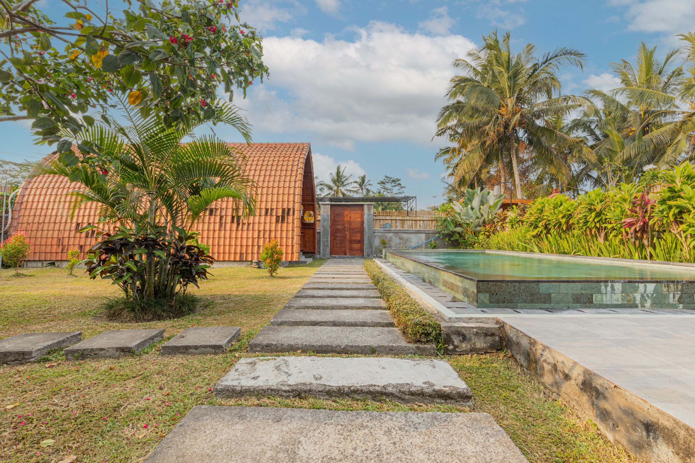 A pathway leading to a building with a tiled roof, surrounded by lush greenery and a swimming pool.