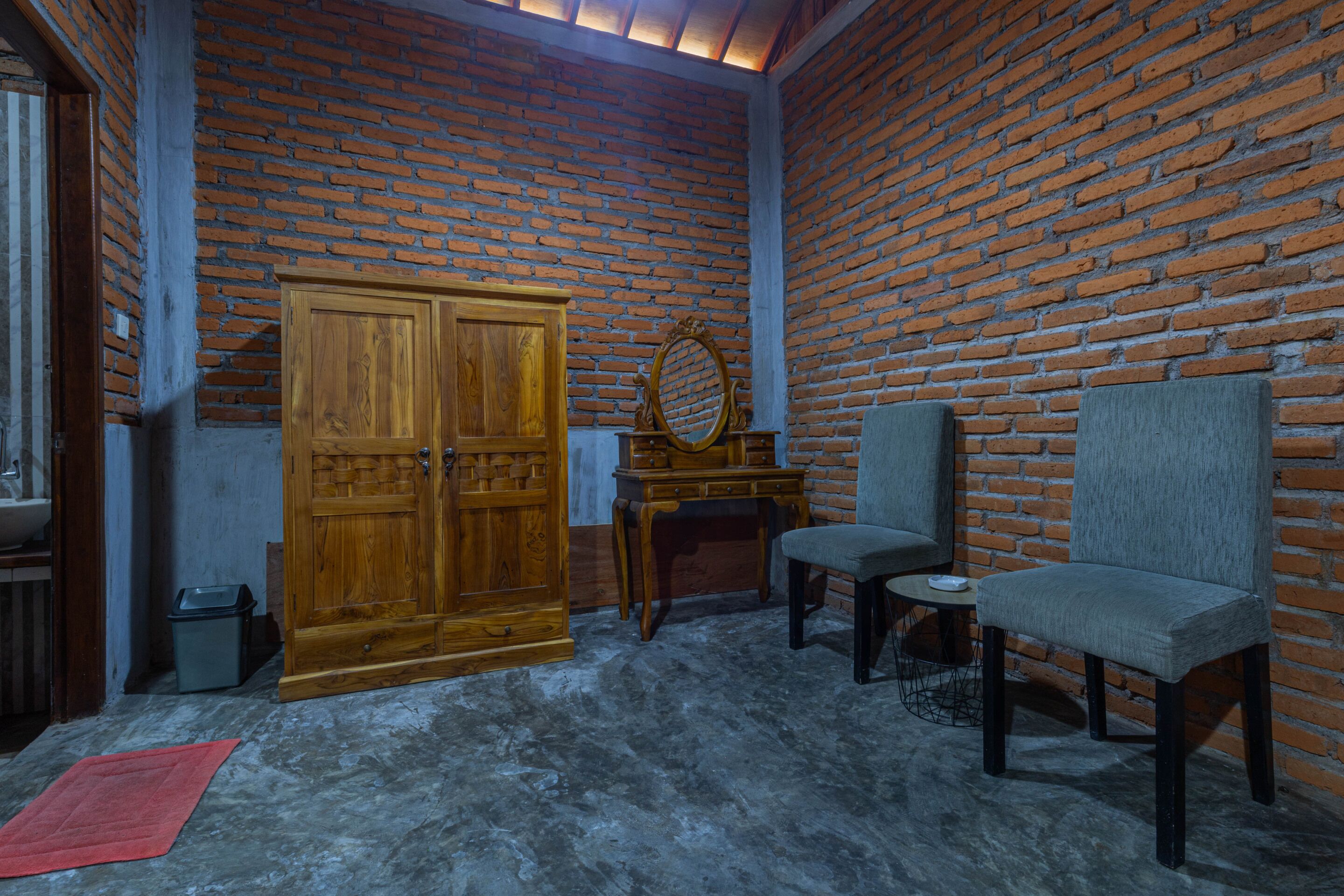 A room with a brick wall, a wooden wardrobe, a small table, two chairs, and a red rug on the floor.