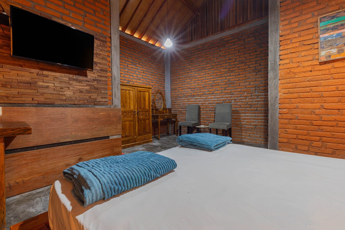 Een kamer met een bakstenen muur, een houten bed, een aan de muur bevestigde televisie en een kleine tafel met een stoel.