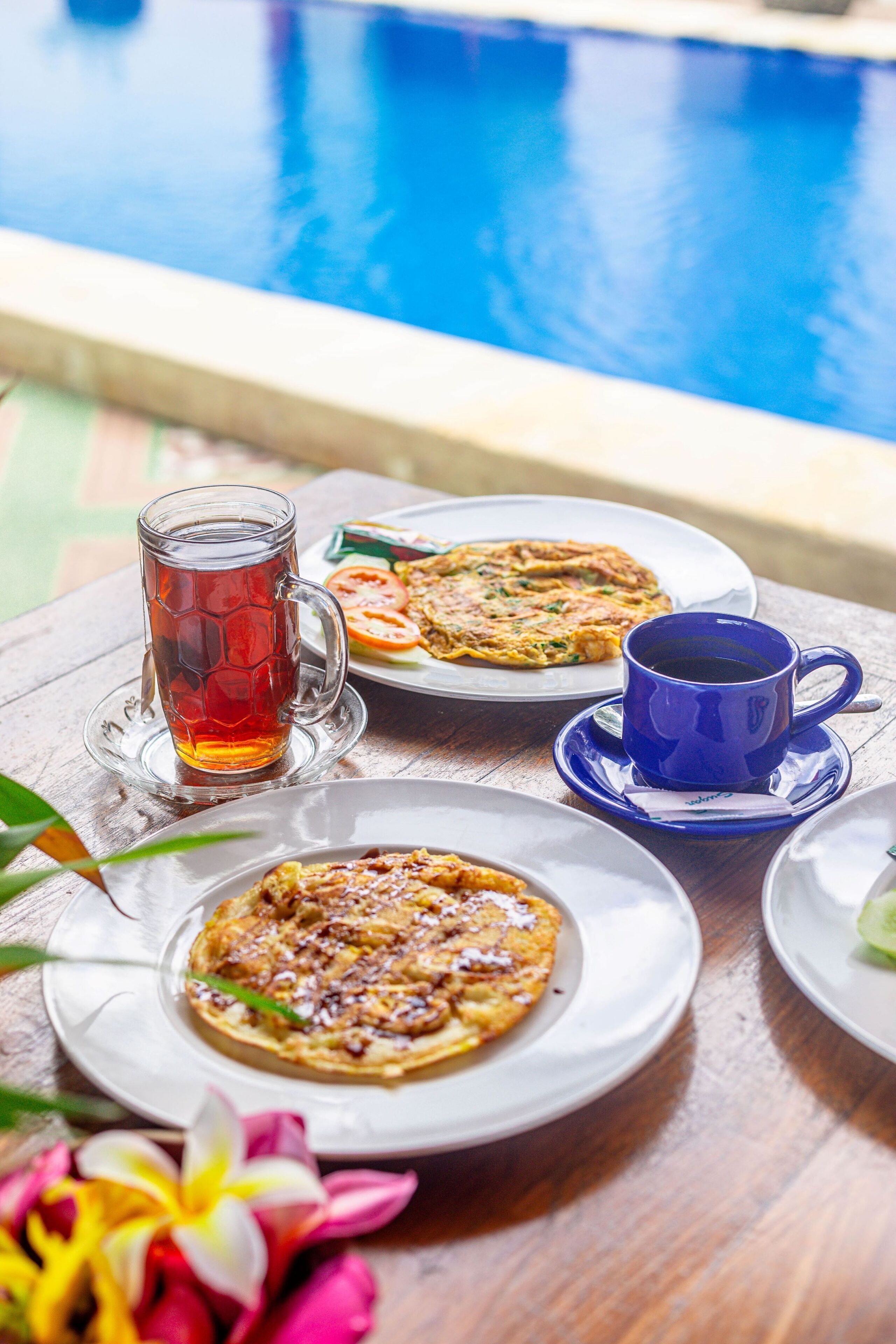 A table with a meal consisting of a drink, a blue cup, and two plates of food near a pool.