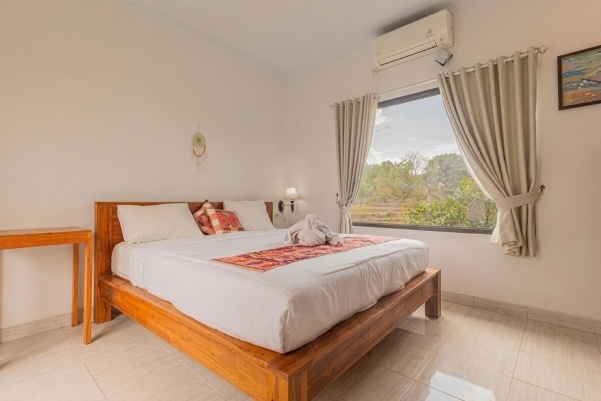 Een slaapkamer met een houten bed, een nachtkastje, een raam met gordijnen en een airconditioning.