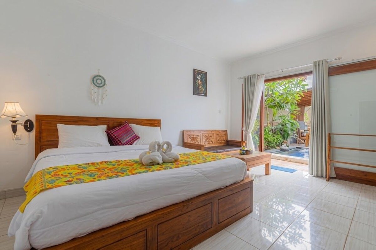 Een hotelkamer met een houten bed, een tafeltje, een bankje en uitzicht op een buitenruimte met planten en een zwembad.
