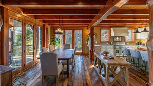 L'intérieur de cette vaste cabane en bois comprend une grande salle à manger avec table et chaises, un coin cuisine et de grandes fenêtres offrant une vue sur la forêt environnante.