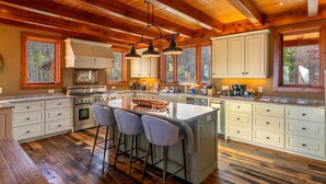 Une cuisine spacieuse dotée d’un grand îlot central, de poutres apparentes en bois au plafond et bénéficiant d’un éclairage naturel abondant grâce à de grandes fenêtres.