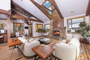 A spacious living room with a fireplace, wooden beams, and large windows.