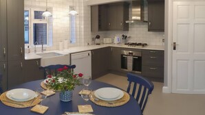 Una cuina moderna amb armaris blancs i grisos fosc, forn integrat i fogó a gas. Hi ha una zona de menjador amb una taula blava preparada per a quatre persones, i un vas amb flors damunt la taula.