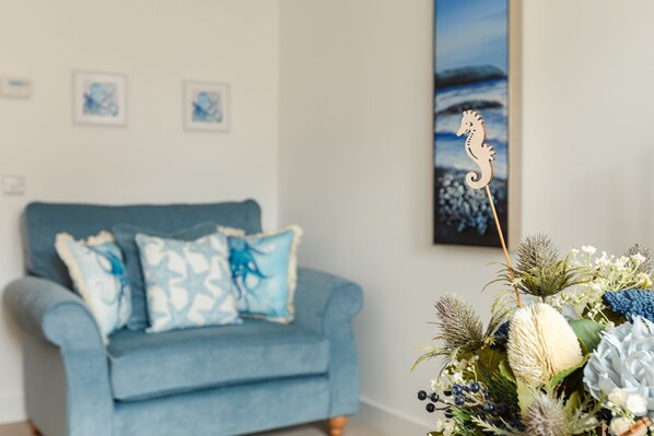 A room with a blue sofa, decorative pillows, a wall-mounted picture, and a wooden seahorse decoration. - Wee Jules By The Sea (Anstruther)