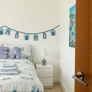 A bedroom with a bed, pillows, a bedside table with a lamp, a wall-mounted string of house-shaped decorations, and a door. - Wee Jules By The Sea (Anstruther)