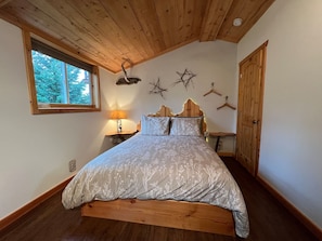 A bedroom with a wooden ceiling, a large window, a bed with a floral bedspread, a bedside table with a lamp, and wall decorations. - Blue Spruce Apartment (Homer)