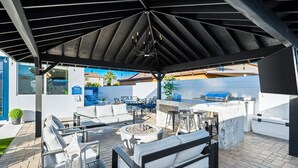 En udendørs terrasse med et overdækket siddeområde, grill og bålsted.