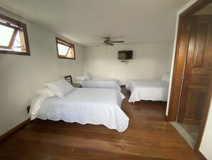 Un dormitorio con dos camas, suelo de madera, ventilador de techo y televisor en la pared.