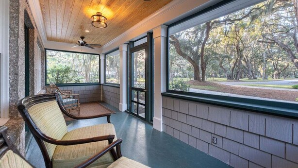 Une véranda avec des fauteuils en osier, un ventilateur de plafond et une vue sur un parc arboré.