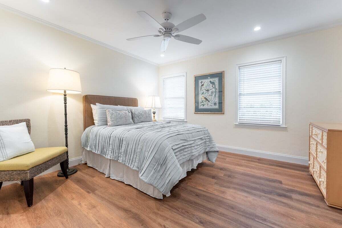 Un dormitorio con cama, una silla, cómoda, ventilador de techo y dos ventanas con persianas.