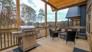 A covered outdoor deck with a barbecue grill, wicker furniture, and a view of trees and houses.
