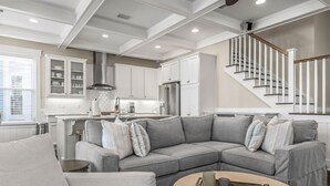 A modern kitchen with white cabinets, a stainless steel refrigerator, and a built-in oven. There is a grey sectional sofa with pillows, a round coffee table, and a staircase with white railings. - Perfect Pitch: Your Serene NatureWalk Haven - 6 Seater Golf Cart (Santa Rosa Beach)