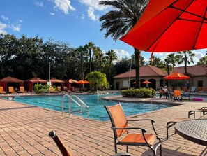 A pool area with lounge chairs and umbrellas, surrounded by palm trees and a building with a tiled roof. - Grand Superior 3BD Townhouse Near Disney (Kissimmee)