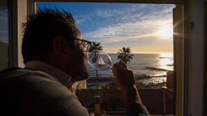 A person looking out a balcony with a view of the ocean and palm trees during sunset.