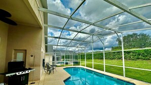 Ein überdachter Pool im Freien mit Grill und Liegestühlen, umgeben von einer Glaseinfassung und Blick auf eine Rasenfläche.