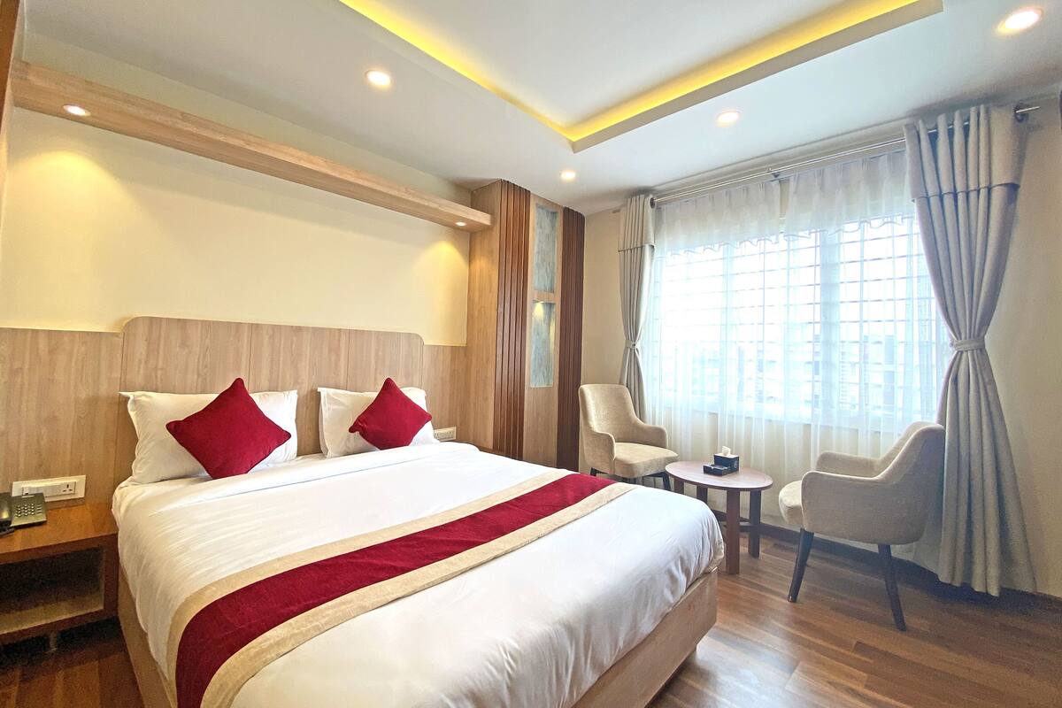 A hotel room with a large bed, a bedside table with a phone, a small table with a lamp, and a chair.