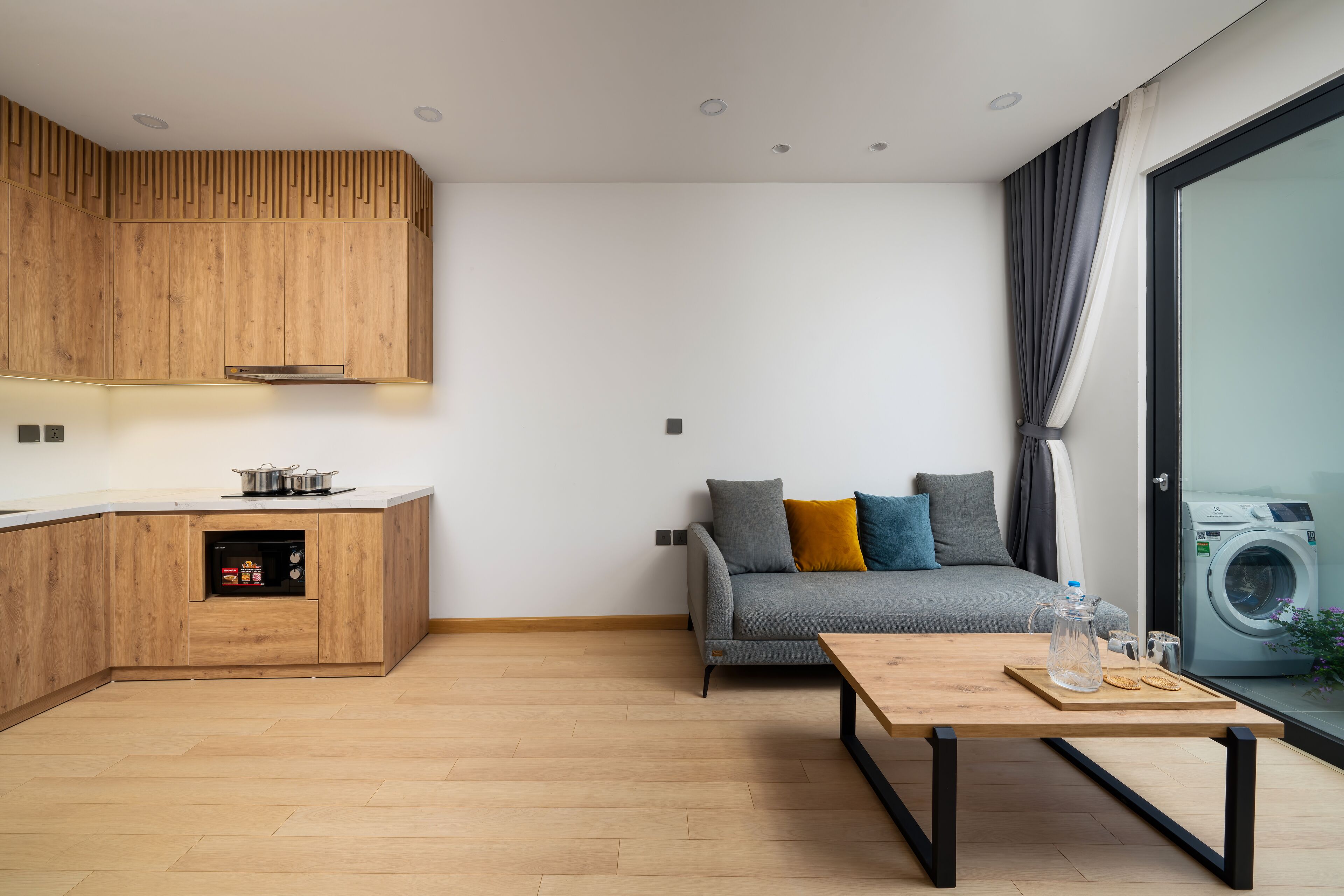 A modern kitchen with wooden cabinets and a built-in oven, a grey sofa with cushions, a wooden coffee table, and a washing machine visible through a glass door.