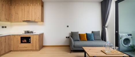 A modern kitchen with wooden cabinets and a built-in oven, a grey sofa with cushions, a wooden coffee table, and a washing machine visible through a glass door.