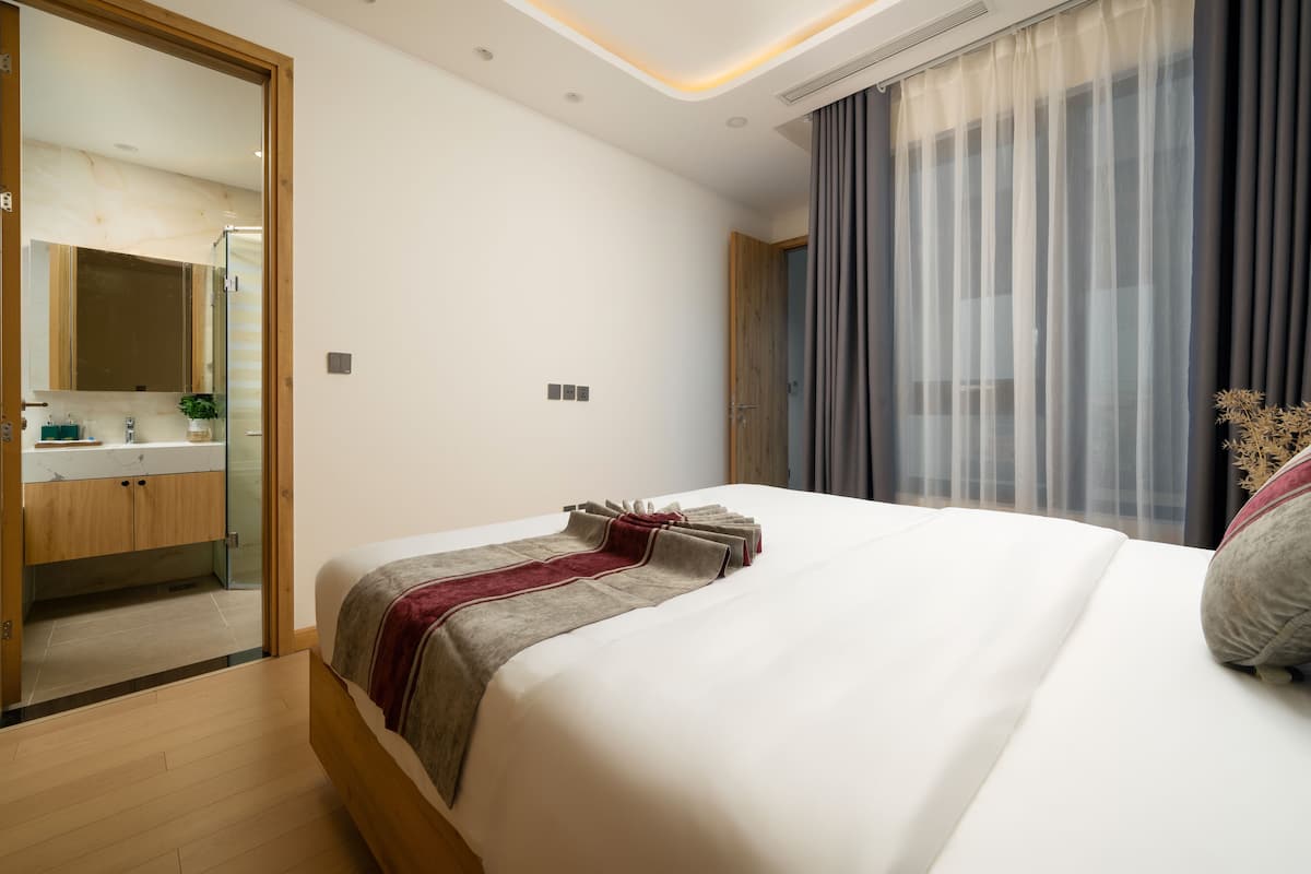 Une chambre d’hôtel moderne avec un grand lit, une salle de bain dotée d’une douche en verre et une vue sur la ville à travers des rideaux transparents.