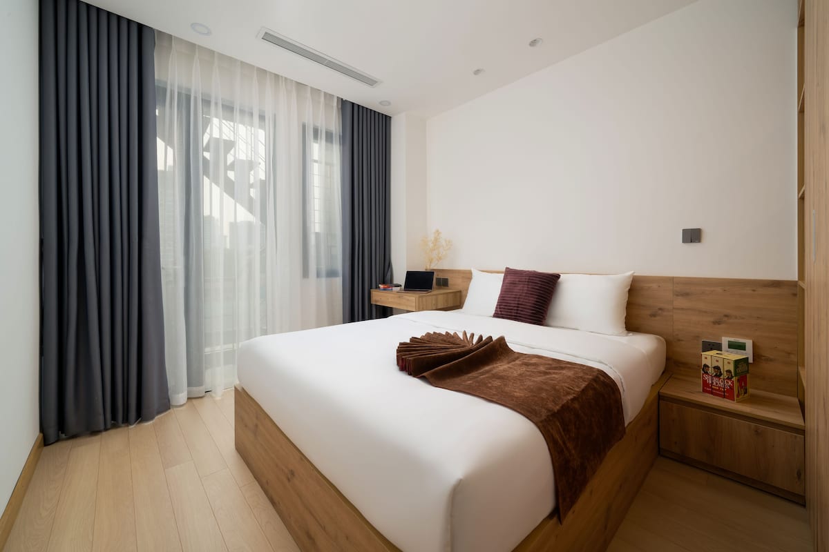 Une chambre d’hôtel moderne avec un grand lit, une tête de lit en bois, une table de chevet et une fenêtre agrémentée de rideaux légers.