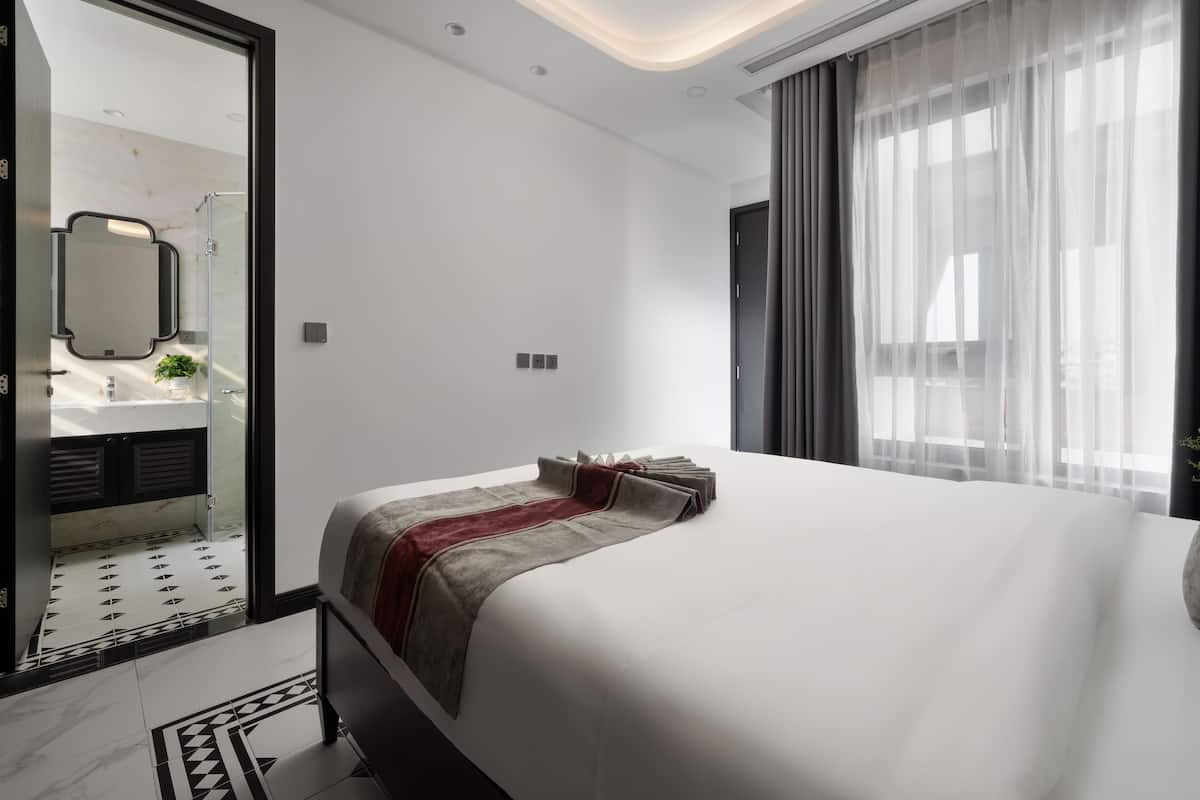 Une chambre d’hôtel moderne avec un grand lit, une salle de bain avec un miroir et une vue sur la ville à travers des rideaux transparents.