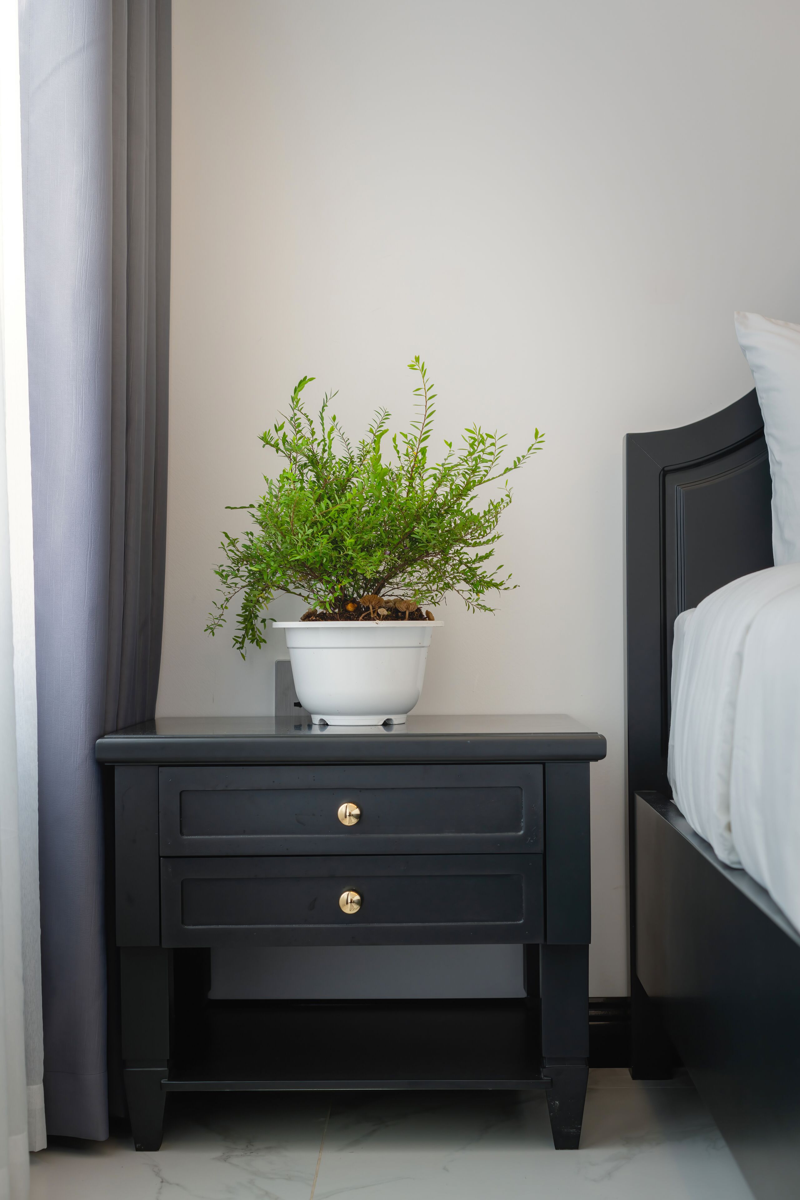 A black nightstand with a white pot containing a green plant, positioned next to a bed with white bedding.