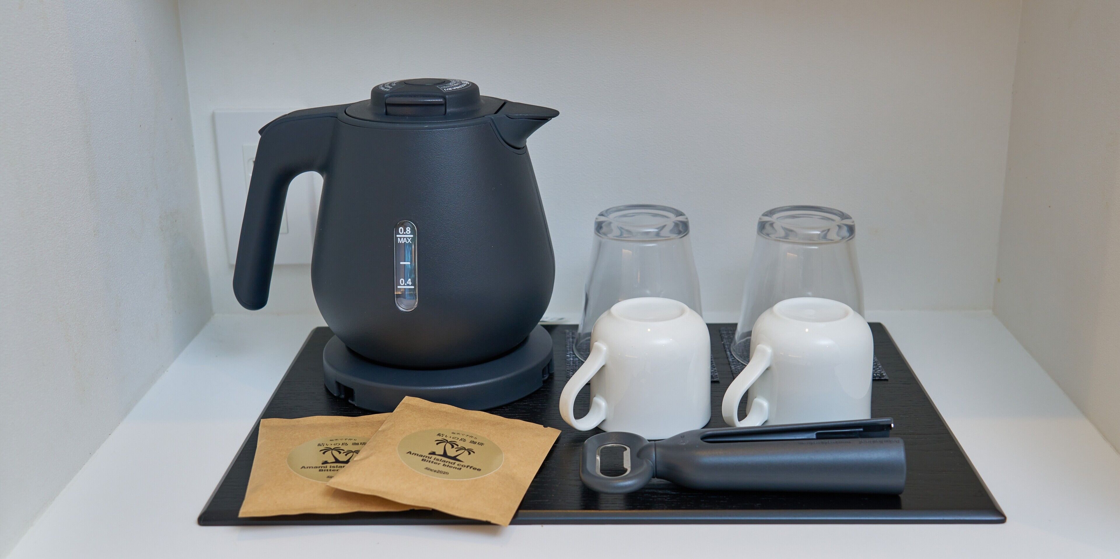 A black electric kettle, two white mugs, two glass tumblers, and two coffee bags on a black tray.