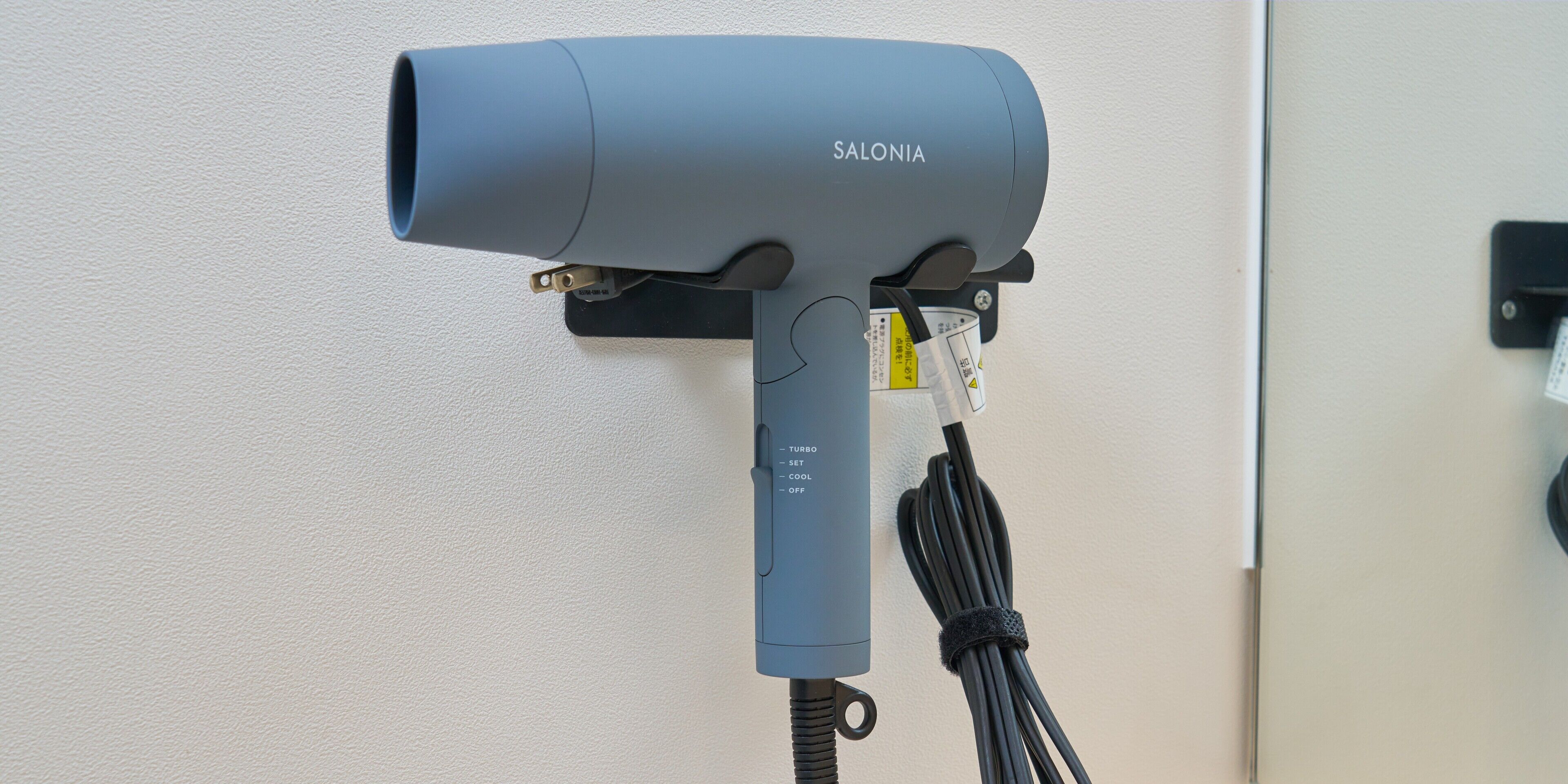 A gray hair dryer with a label "SALONIA" mounted on a wall, connected to a power outlet.
