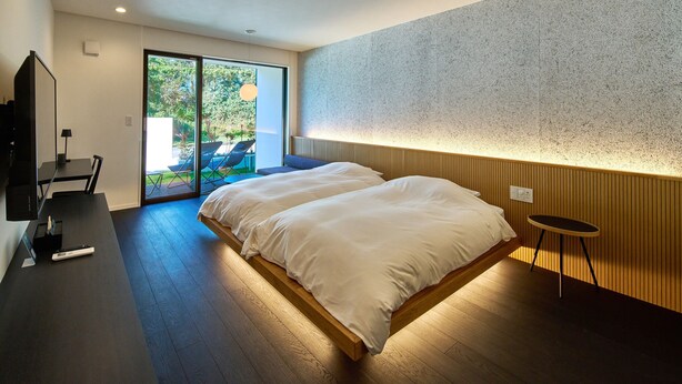 A modern bedroom with a large bed, a desk with a computer, and a sliding glass door leading to an outdoor area with chairs. - LUXE TECH VILLA ASHITOKU (Tatsugo)