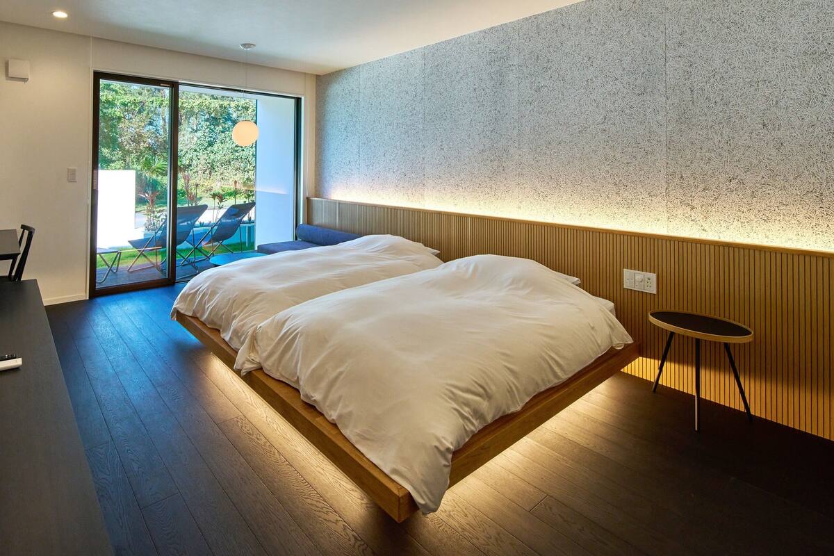 Un dormitorio moderno con una cama grande, un escritorio con ordenador y una puerta corrediza de cristal que da a un espacio exterior con sillas.