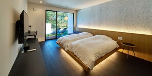 A modern bedroom with a large bed, a desk with a computer, and a sliding glass door leading to an outdoor area with chairs. - LUXE TECH VILLA ASHITOKU (Tatsugo)