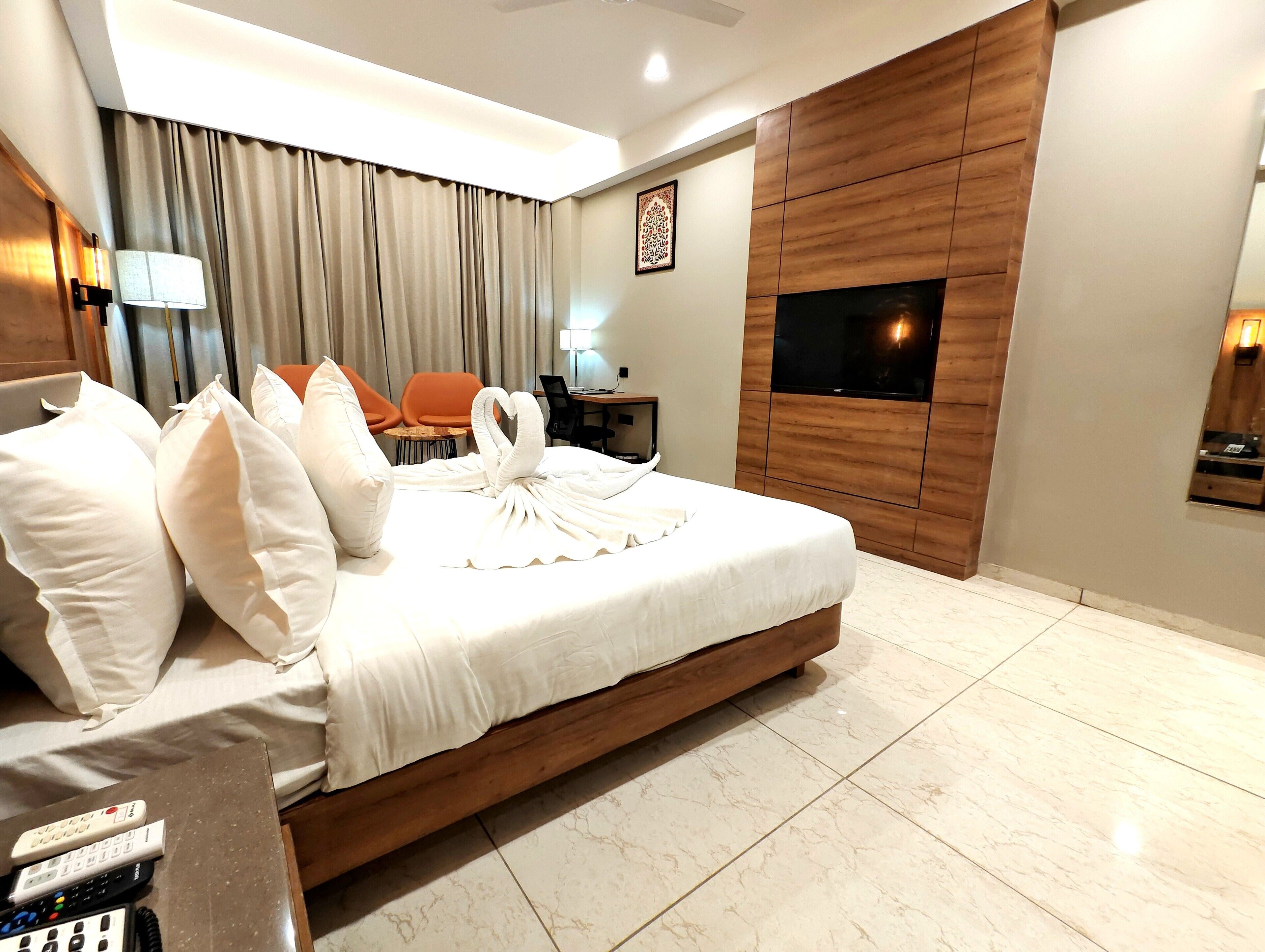 A hotel room with a large bed, a TV mounted on the wall, a desk with a chair, and a small table with a lamp.