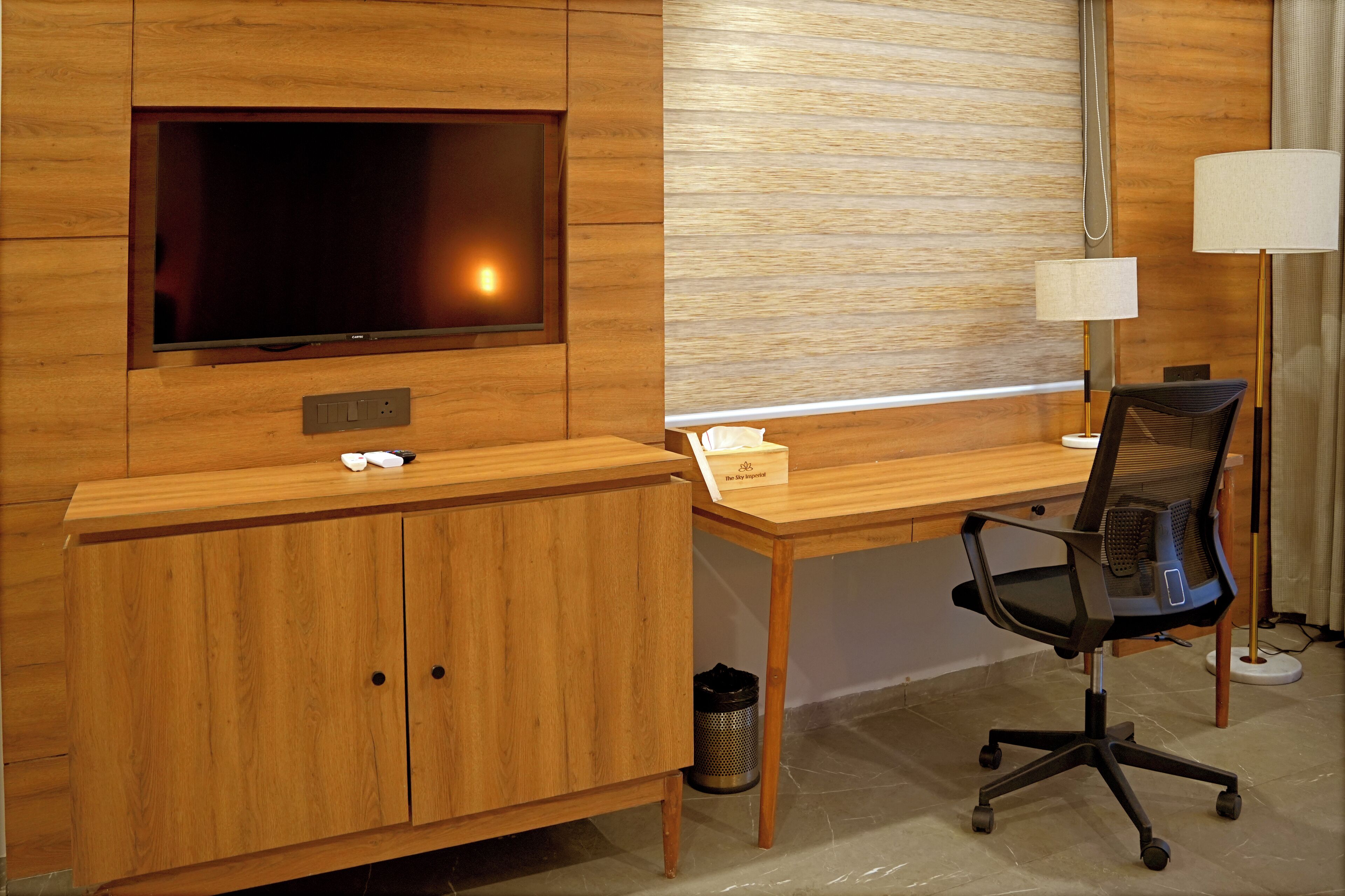 Ein modernes Hotelzimmer mit Schreibtisch aus Holz, einem an der Wand montierten Fernseher, einer Lampe und einem Papierkorb.
