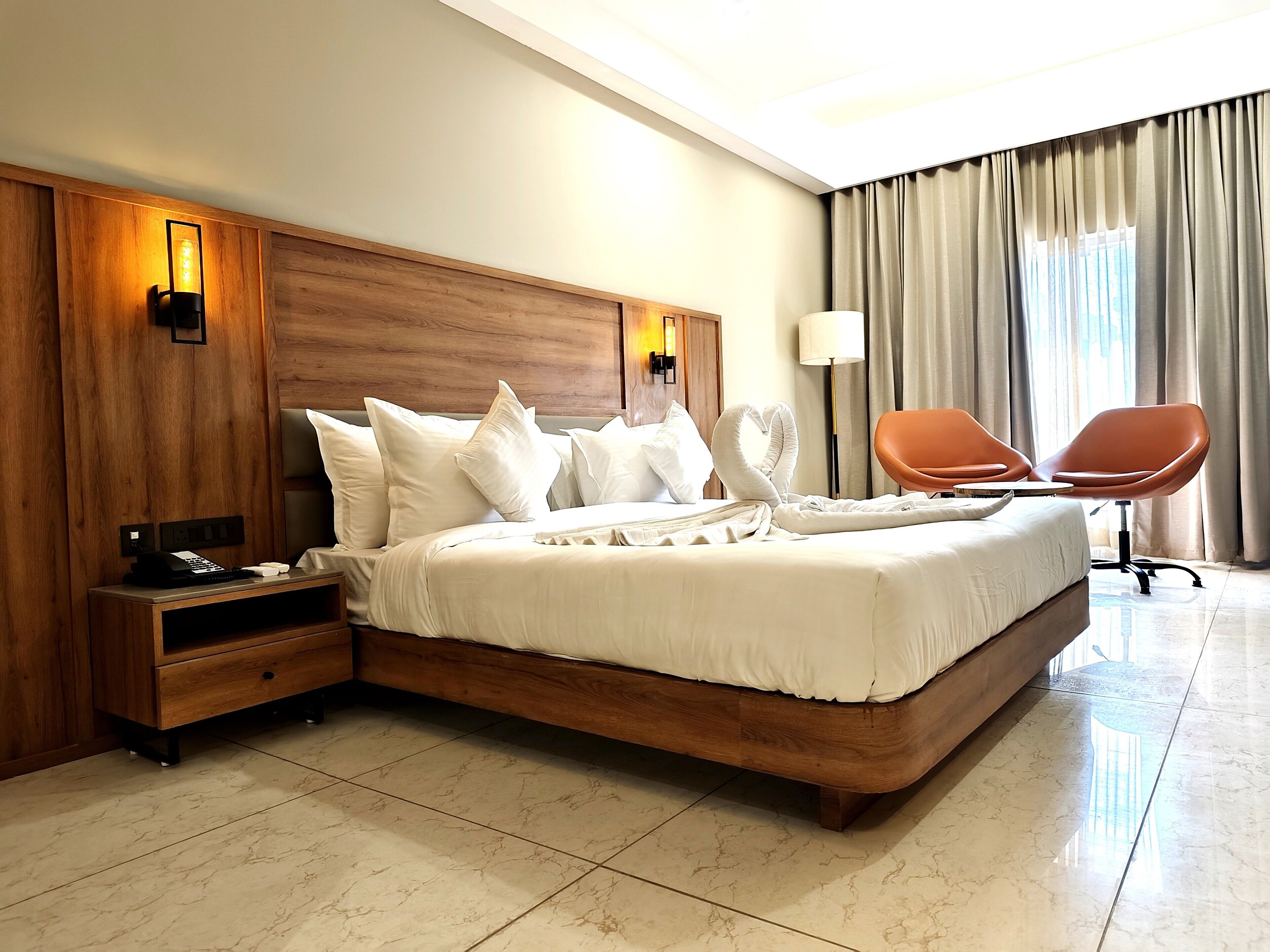 A hotel room with a large bed, wooden headboard, bedside table with phone and charger, and a chair.
