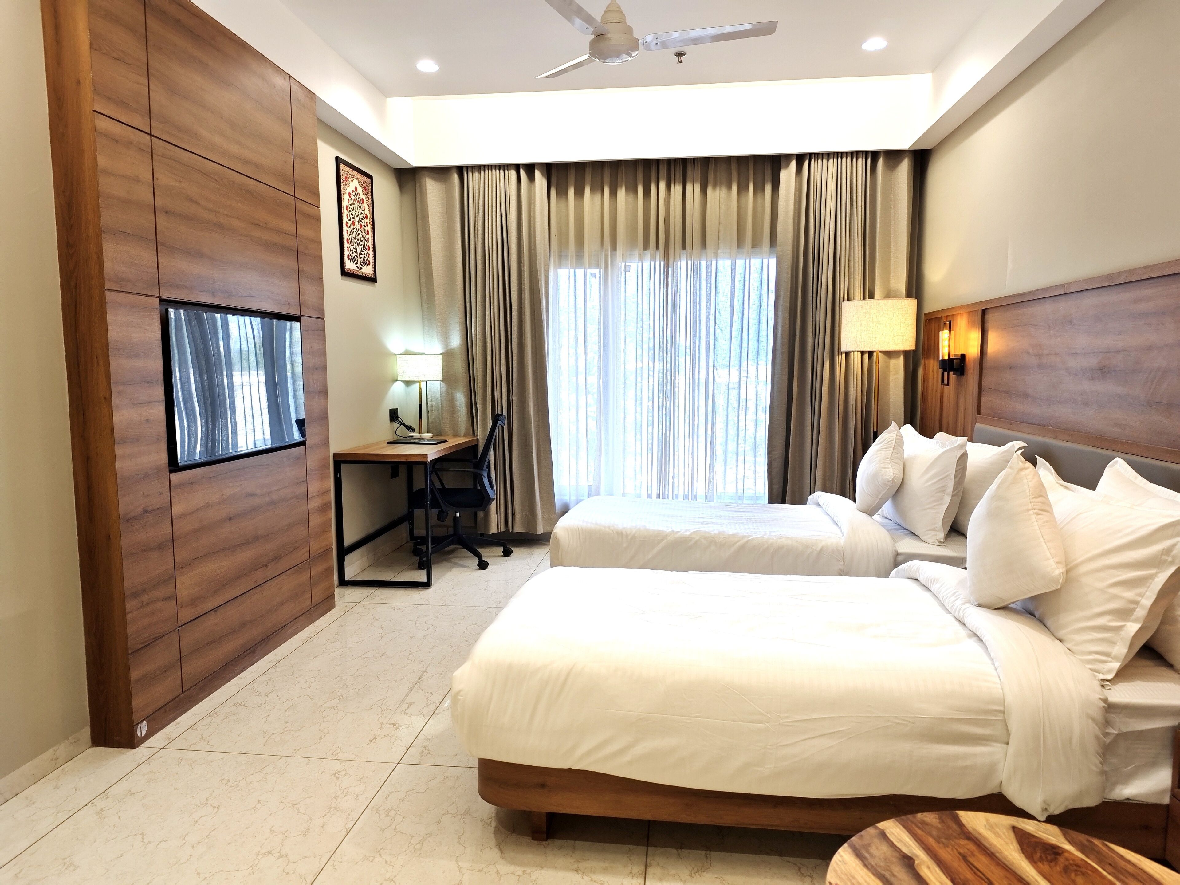 A hotel room with two beds, a desk, a chair, a TV, and a ceiling fan.