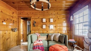 L’intérieur chaleureux d’un chalet, avec un décor en bois, un canapé vert, un petit pouf orange et une cheminée en pierre.
