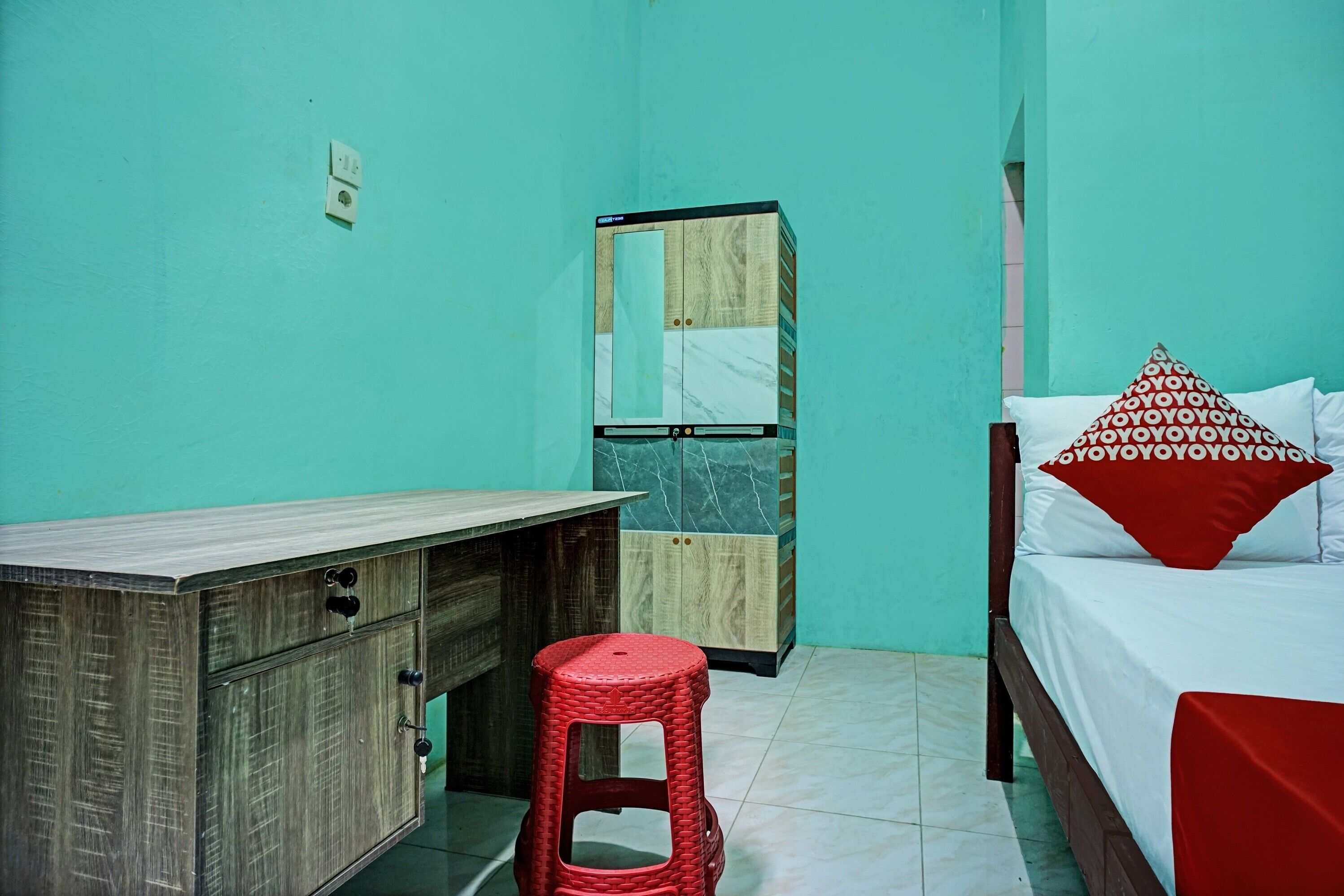 A small, well-lit room with a wooden desk, a red stool, a wardrobe, and a bed with patterned pillows.