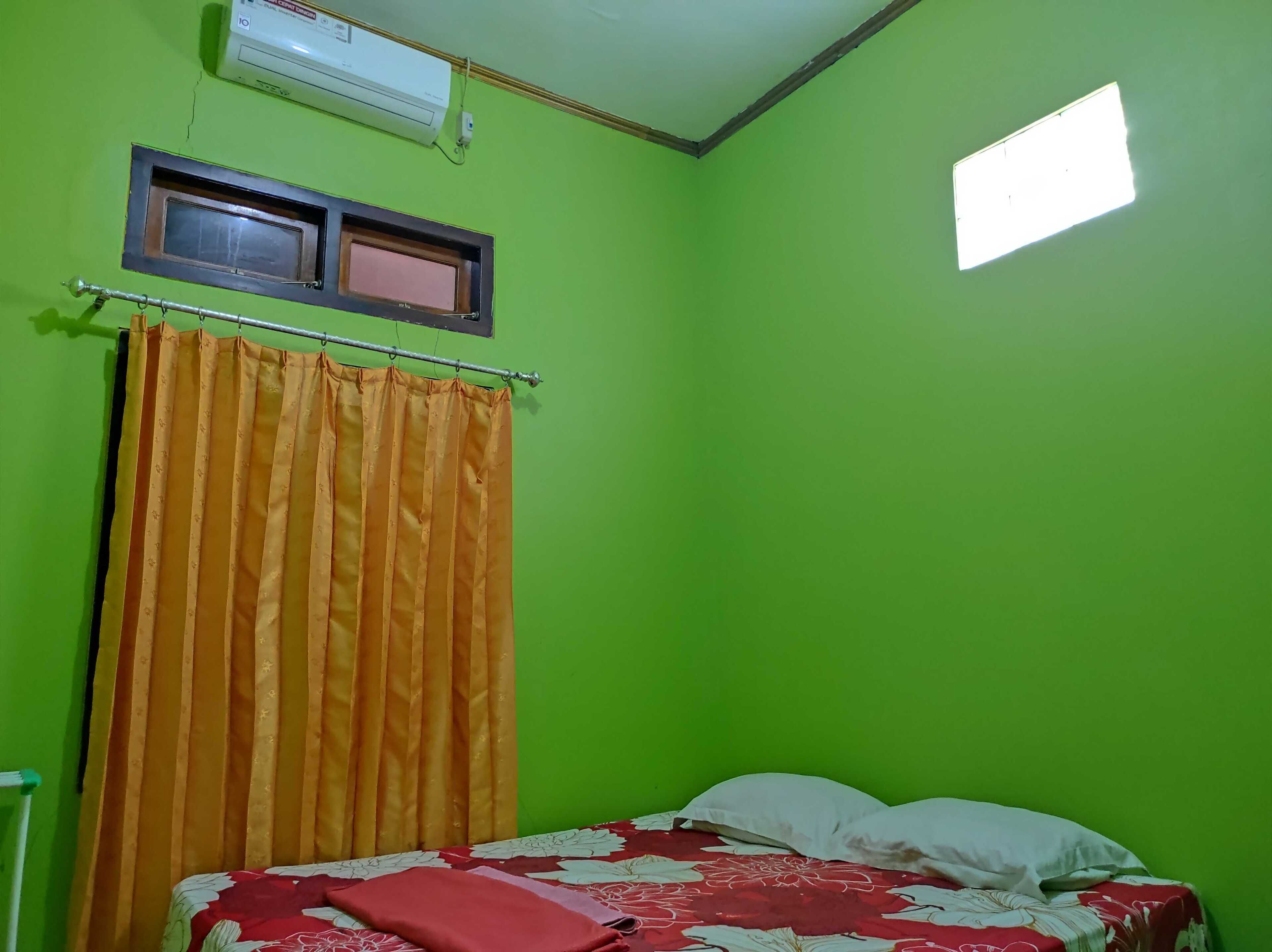A room with green walls, a bed with a floral bedspread, an air conditioner, a window with a blind, and a curtain.