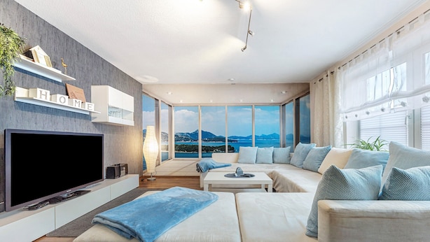 A modern living room with a large flat-screen TV, a sofa, a coffee table, and a view of the sea.